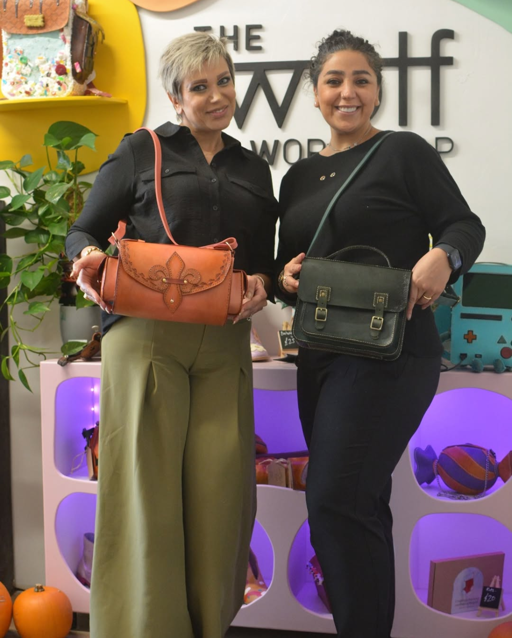 Photo of two women standing side by side inside a boutique or craft shop, smiling at the camera while modeling handmade shoulder bags. The woman on the left has short blonde hair and wears a black button-up shirt with loose olive-green pants, holding a brown leather handbag with decorative stitched detailing and a coral strap. The woman on the right has dark curly hair tied back and wears a black long-sleeve outfit, carrying a structured dark green handbag with buckle accents and a matching shoulder strap. Behind them are colorful shelves displaying bags, small accessories, plants, and decorative items, with soft purple accent lighting that creates a warm, stylish retail setting.