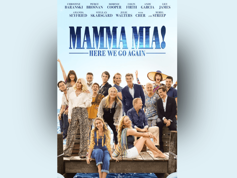 Poster for Mamma Mia! Here We Go Again featuring a large ensemble cast gathered together on a wooden dock above bright blue water. At the center, a smiling blonde woman in dark overalls stands surrounded by friends and family members dressed in colorful summer clothing, including patterned dresses, light shirts, and suits. Two younger women sit casually at the front of the dock, while the rest of the group stands behind them in relaxed, cheerful poses. The pale blue background gives the poster a bright, airy feel, with large bold blue text at the top reading Mamma Mia! Here We Go Again! The overall composition conveys a fun, celebratory atmosphere centered around music, friendship, and family reunion.