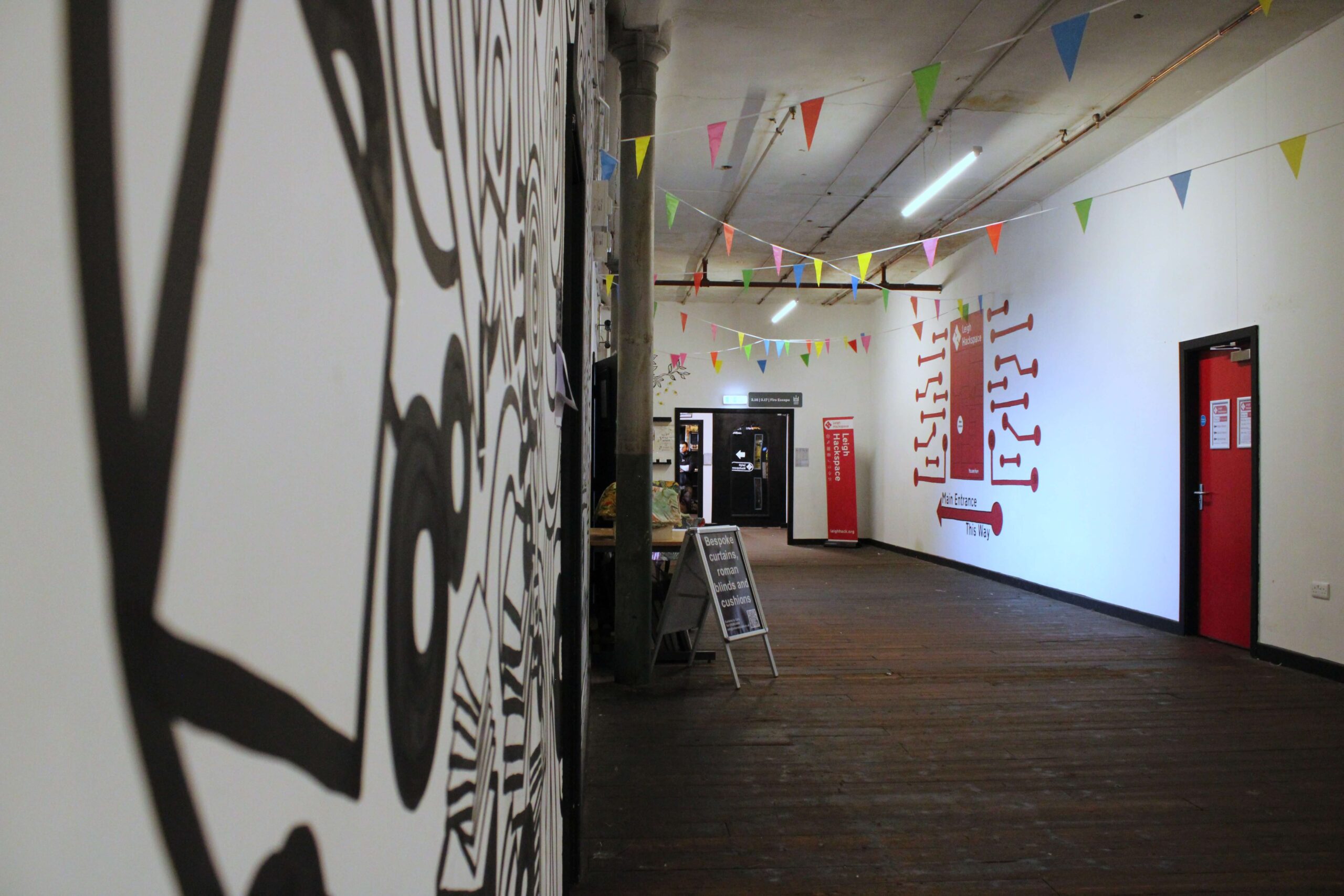 Long corridor inside Spinners Mill featuring original wooden flooring, colourful bunting, wall artwork, and wayfinding signage for businesses and facilities.