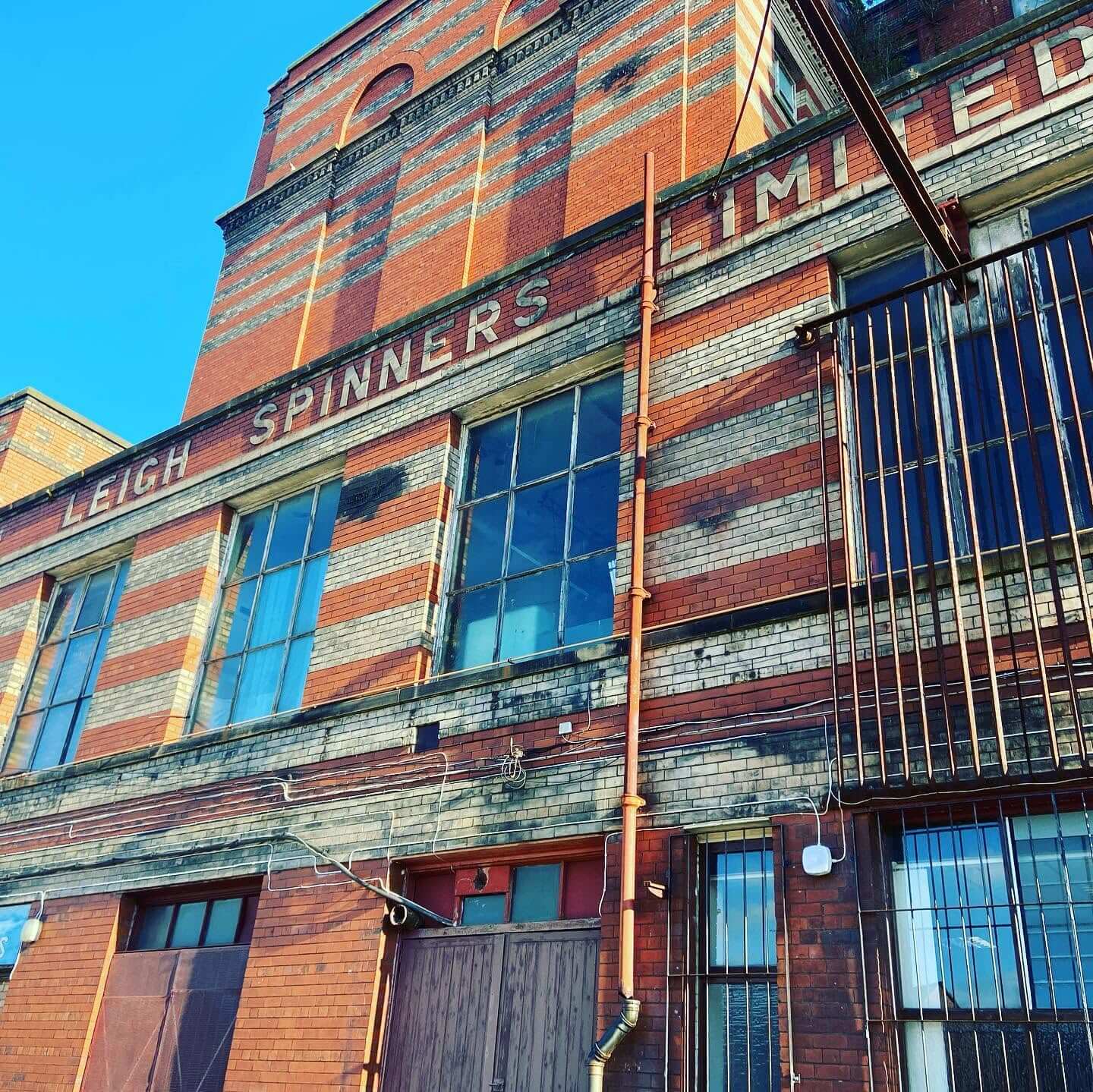 Exterior view of Leigh Spinners Mill showing the historic red brick façade, large mill windows and the original Leigh Spinners Mill lettering on the building.